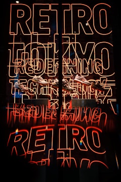 Bright neon sign featuring the words 'RETRO TOKYO TECH' in orange-red hues, with a reflection creating a mirrored effect. Below the neon, several sneakers are displayed.