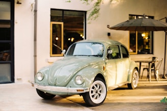A vintage green Volkswagen Beetle car is parked in an outdoor setting with a patio area. The background features a light-colored building with windows and a large patio umbrella shading a table and several chairs. The setting appears calm and relaxed, perhaps part of a cafe or restaurant environment.
