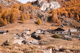 A serene mountain village landscape featuring rustic stone houses nestled among golden autumn trees. A rugged path leads through the scene, where a group of hikers walks toward the village. The backdrop includes rocky cliffs and a clear sky, adding to the tranquility of the setting.
