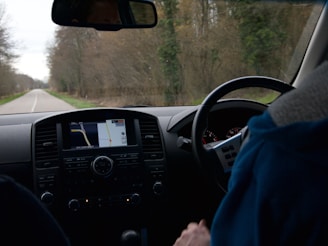 The image depicts the interior of a moving vehicle, showing the dashboard with a navigation screen displaying a map. A person wearing a blue jacket is holding the steering wheel. Outside the car, a rural road lined with trees is visible, suggesting a drive through a wooded area.