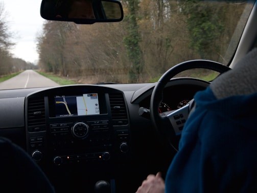 The image depicts the interior of a moving vehicle, showing the dashboard with a navigation screen displaying a map. A person wearing a blue jacket is holding the steering wheel. Outside the car, a rural road lined with trees is visible, suggesting a drive through a wooded area.