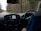 The image depicts the interior of a moving vehicle, showing the dashboard with a navigation screen displaying a map. A person wearing a blue jacket is holding the steering wheel. Outside the car, a rural road lined with trees is visible, suggesting a drive through a wooded area.