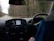 The image depicts the interior of a moving vehicle, showing the dashboard with a navigation screen displaying a map. A person wearing a blue jacket is holding the steering wheel. Outside the car, a rural road lined with trees is visible, suggesting a drive through a wooded area.