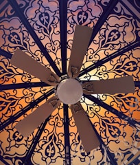 A ceiling fan is viewed from below, surrounded by an intricate and ornate pattern. The pattern features floral and geometric designs in dark lines against a warm, glowing background that transitions from orange to pink hues.