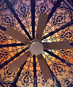 A ceiling fan is viewed from below, surrounded by an intricate and ornate pattern. The pattern features floral and geometric designs in dark lines against a warm, glowing background that transitions from orange to pink hues.
