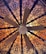 A ceiling fan is viewed from below, surrounded by an intricate and ornate pattern. The pattern features floral and geometric designs in dark lines against a warm, glowing background that transitions from orange to pink hues.