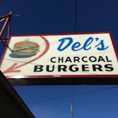 A rectangular outdoor sign with a red border showcasing the name 'Del's Charcoal Burgers' in bold black and blue text. An illustration of a hamburger with a red arrow pointing towards it is featured on the left side of the sign. The background is a clear blue sky.
