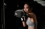 A woman in athletic wear is intensely focused as she throws a punch, wearing boxing gloves. Her stance and expression suggest concentration and determination. The background is dark, emphasizing her figure and action.