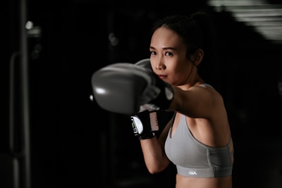 A woman in athletic wear is intensely focused as she throws a punch, wearing boxing gloves. Her stance and expression suggest concentration and determination. The background is dark, emphasizing her figure and action.