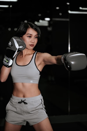 A woman wearing sports attire is engaged in a boxing stance in a dimly lit gym or studio. She is wearing boxing gloves and looks focused on her activity.