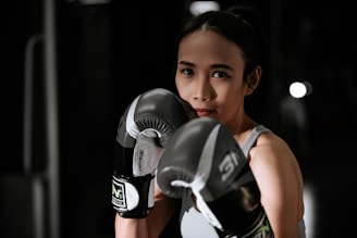 A woman wearing boxing gloves is poised in a fighting stance. Her expression is focused and determined, and she is standing in a dimly lit space, possibly a gym.