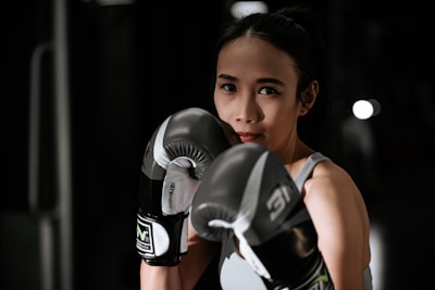 A woman wearing boxing gloves is poised in a fighting stance. Her expression is focused and determined, and she is standing in a dimly lit space, possibly a gym.