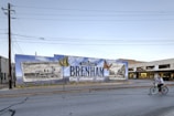 A large mural on a wall promotes Downtown Brenham, Texas, featuring illustrations of a vintage train and a historic building alongside bright orange butterflies. The wall is painted with a mix of blue and green, and the mural includes the words 'Cool. Authentic. Texas.' A man rides a bicycle in front of the mural down the street. The scene is set in a small town environment with clear skies.