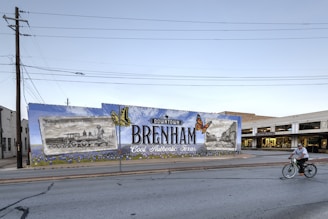 A large mural on a wall promotes Downtown Brenham, Texas, featuring illustrations of a vintage train and a historic building alongside bright orange butterflies. The wall is painted with a mix of blue and green, and the mural includes the words 'Cool. Authentic. Texas.' A man rides a bicycle in front of the mural down the street. The scene is set in a small town environment with clear skies.