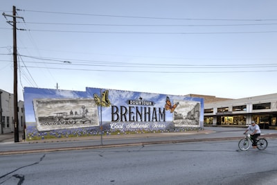 A large mural on a wall promotes Downtown Brenham, Texas, featuring illustrations of a vintage train and a historic building alongside bright orange butterflies. The wall is painted with a mix of blue and green, and the mural includes the words 'Cool. Authentic. Texas.' A man rides a bicycle in front of the mural down the street. The scene is set in a small town environment with clear skies.
