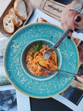 A bowl of pasta is elegantly presented on a vibrant turquoise plate, garnished with basil and a sprinkling of cheese. Slices of bread rest on a wooden platter beside the main dish. A hand with dark nail polish and silver rings is holding a fork, ready to dive into the meal. There are papers or magazines laid beneath the plate, adding a casual dining vibe.