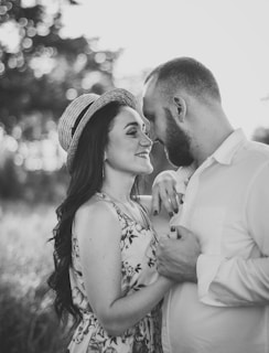 A monochrome photograph of a couple standing close together in a field. The woman is wearing a floral dress and a straw hat, while the man is in a light-colored shirt. They are facing each other, holding hands, and smiling softly with their foreheads touching.