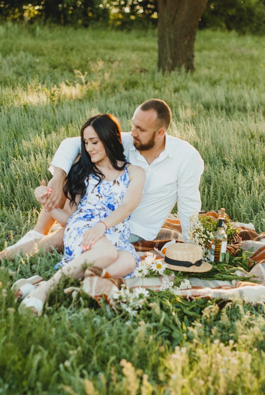 A couple sits together on a picnic blanket in a lush, green field. The woman is wearing a white and blue floral dress, while the man is dressed in a white shirt and light-colored pants. They appear relaxed and are surrounded by various picnic items, including a straw hat, a bottle, and flowers. The setting is serene, with soft light filtering through the trees in the background.