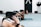 Several individuals are practicing yoga in a studio setting. They are on mats in a pose that involves kneeling and leaning forward with arms stretched out. The focus is on the foreground participant with tattoos, and there's a punching bag and closed door visible in the background.