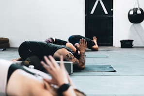Several individuals are practicing yoga in a studio setting. They are on mats in a pose that involves kneeling and leaning forward with arms stretched out. The focus is on the foreground participant with tattoos, and there's a punching bag and closed door visible in the background.