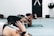 Several individuals are practicing yoga in a studio setting. They are on mats in a pose that involves kneeling and leaning forward with arms stretched out. The focus is on the foreground participant with tattoos, and there's a punching bag and closed door visible in the background.