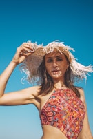 A woman in a colorful patterned swimsuit holds a wide-brimmed, straw hat against a clear blue sky. The hat casts intricate shadows across her face, highlighting her features. Her hair is dark and flowing, and her expression is relaxed.