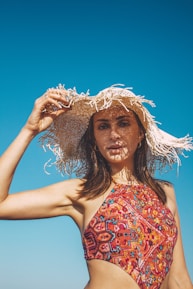A woman in a colorful patterned swimsuit holds a wide-brimmed, straw hat against a clear blue sky. The hat casts intricate shadows across her face, highlighting her features. Her hair is dark and flowing, and her expression is relaxed.