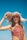 A woman in a colorful patterned swimsuit holds a wide-brimmed, straw hat against a clear blue sky. The hat casts intricate shadows across her face, highlighting her features. Her hair is dark and flowing, and her expression is relaxed.