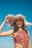 A woman in a colorful patterned swimsuit holds a wide-brimmed, straw hat against a clear blue sky. The hat casts intricate shadows across her face, highlighting her features. Her hair is dark and flowing, and her expression is relaxed.