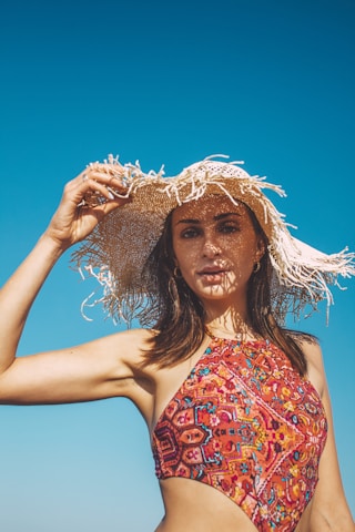 A woman in a colorful patterned swimsuit holds a wide-brimmed, straw hat against a clear blue sky. The hat casts intricate shadows across her face, highlighting her features. Her hair is dark and flowing, and her expression is relaxed.