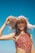 A woman in a colorful patterned swimsuit holds a wide-brimmed, straw hat against a clear blue sky. The hat casts intricate shadows across her face, highlighting her features. Her hair is dark and flowing, and her expression is relaxed.