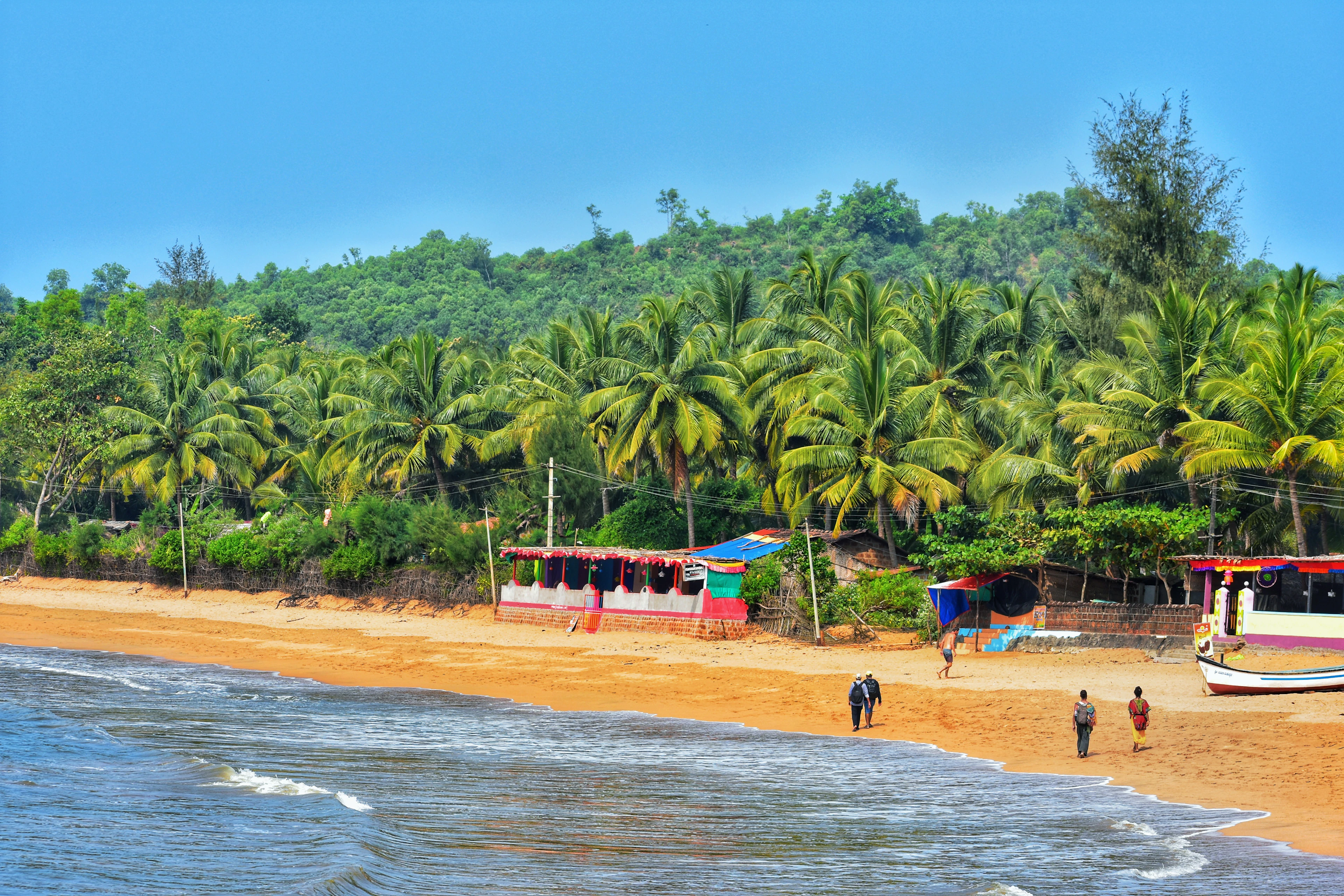 A palm-fringed golden-sand beach with colorful shacks, fishing boats and people walking along the shore.