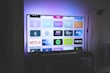 A flat-screen television is displayed in a dimly lit room, showcasing a variety of streaming service apps on its screen, such as Netflix, Prime Video, and Spotify. The TV is positioned on a black stand beside a tall floor lamp, casting a soft ambient light on the surrounding area.