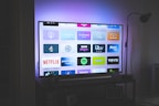 A flat-screen television is displayed in a dimly lit room, showcasing a variety of streaming service apps on its screen, such as Netflix, Prime Video, and Spotify. The TV is positioned on a black stand beside a tall floor lamp, casting a soft ambient light on the surrounding area.