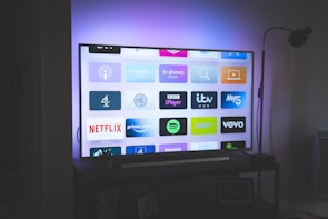 A flat-screen television is displayed in a dimly lit room, showcasing a variety of streaming service apps on its screen, such as Netflix, Prime Video, and Spotify. The TV is positioned on a black stand beside a tall floor lamp, casting a soft ambient light on the surrounding area.
