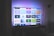 A flat-screen television is displayed in a dimly lit room, showcasing a variety of streaming service apps on its screen, such as Netflix, Prime Video, and Spotify. The TV is positioned on a black stand beside a tall floor lamp, casting a soft ambient light on the surrounding area.