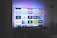 A flat-screen television is displayed in a dimly lit room, showcasing a variety of streaming service apps on its screen, such as Netflix, Prime Video, and Spotify. The TV is positioned on a black stand beside a tall floor lamp, casting a soft ambient light on the surrounding area.