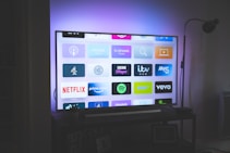 A flat-screen television is displayed in a dimly lit room, showcasing a variety of streaming service apps on its screen, such as Netflix, Prime Video, and Spotify. The TV is positioned on a black stand beside a tall floor lamp, casting a soft ambient light on the surrounding area.