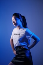 A person stands against a gradient blue background, looking towards the light. The individual is wearing a white T-shirt with 'Metallica' written on it and a dark leather skirt. The lighting creates a dramatic effect with blue tones highlighting the facial features and clothing.