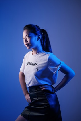 A person stands against a gradient blue background, looking towards the light. The individual is wearing a white T-shirt with 'Metallica' written on it and a dark leather skirt. The lighting creates a dramatic effect with blue tones highlighting the facial features and clothing.