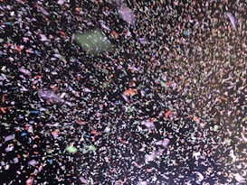 A vibrant and chaotic scene filled with multicolored confetti scattered against a dark background. Tiny pieces of paper in colors like red, green, white, and blue create a dynamic and festive atmosphere.