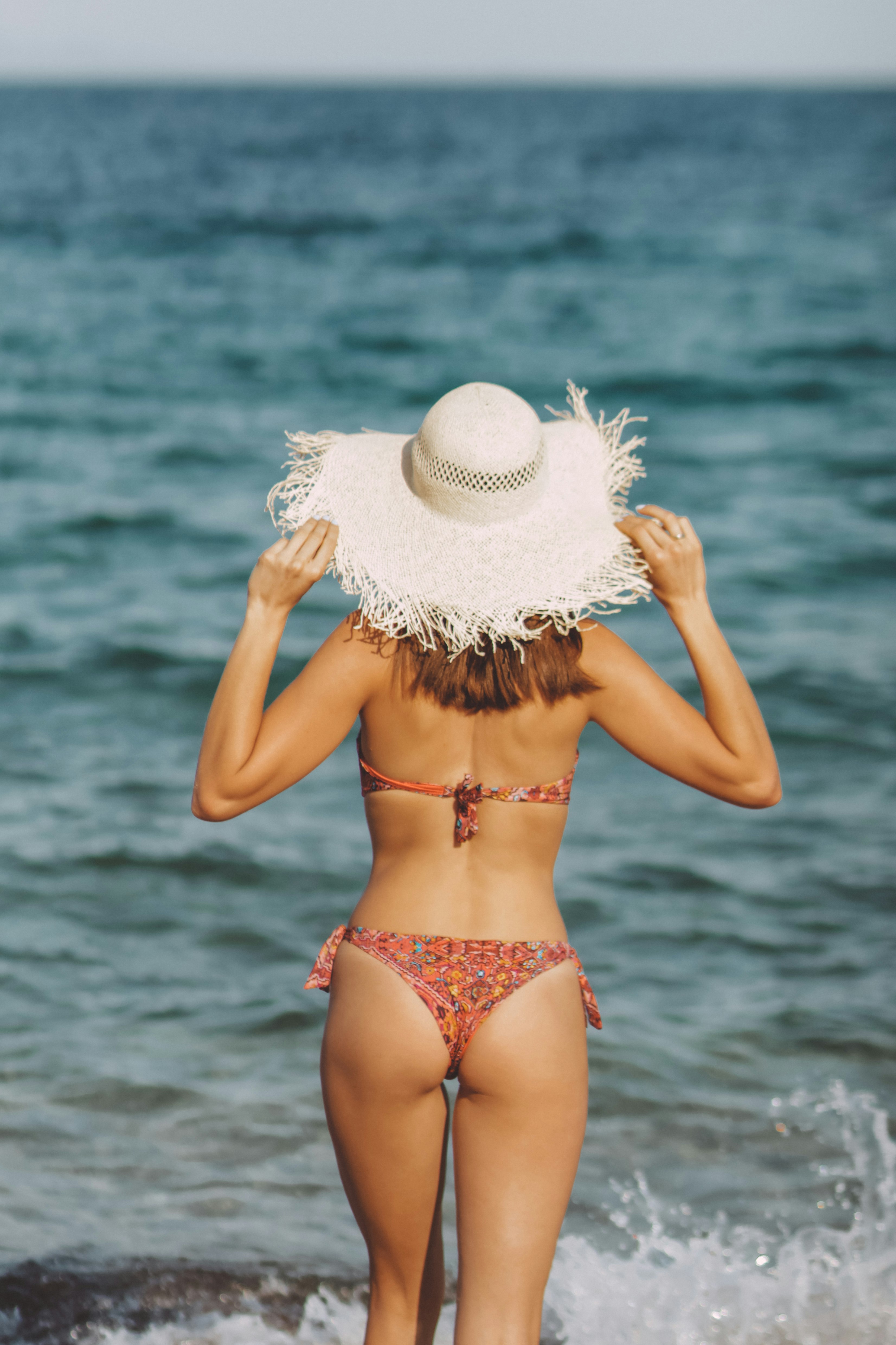 woman in white and red floral bikini wearing white sun hat standing on sea shore during