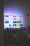 A flat-screen television is displaying a variety of streaming app icons, including Netflix, Prime Video, Spotify, and ITV Hub among others. The TV is placed on a media console with a small potted plant beside it. A tall lamp stands to the right, and a faint, ambient light casts a purple hue on the wall above the TV.