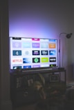 A flat-screen television is displaying a variety of streaming app icons, including Netflix, Prime Video, Spotify, and ITV Hub among others. The TV is placed on a media console with a small potted plant beside it. A tall lamp stands to the right, and a faint, ambient light casts a purple hue on the wall above the TV.