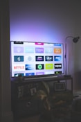 A flat-screen television is displaying a variety of streaming app icons, including Netflix, Prime Video, Spotify, and ITV Hub among others. The TV is placed on a media console with a small potted plant beside it. A tall lamp stands to the right, and a faint, ambient light casts a purple hue on the wall above the TV.