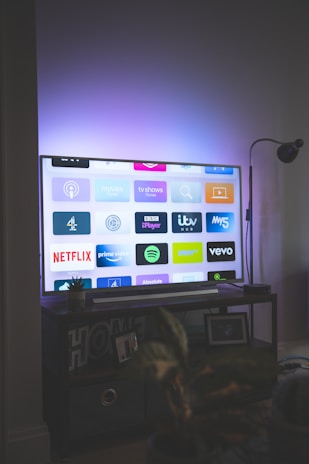 A flat-screen television is displaying a variety of streaming app icons, including Netflix, Prime Video, Spotify, and ITV Hub among others. The TV is placed on a media console with a small potted plant beside it. A tall lamp stands to the right, and a faint, ambient light casts a purple hue on the wall above the TV.