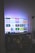 A flat-screen television is displaying a variety of streaming app icons, including Netflix, Prime Video, Spotify, and ITV Hub among others. The TV is placed on a media console with a small potted plant beside it. A tall lamp stands to the right, and a faint, ambient light casts a purple hue on the wall above the TV.