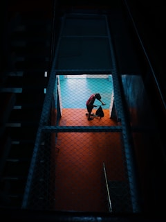 A person in a red shirt is mopping or sweeping a brightly lit floor, viewed through a fenced or grated enclosure, creating a contrast with the dark surrounding area.
