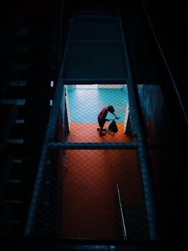 A person in a red shirt is mopping or sweeping a brightly lit floor, viewed through a fenced or grated enclosure, creating a contrast with the dark surrounding area.