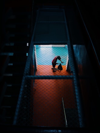 A person in a red shirt is mopping or sweeping a brightly lit floor, viewed through a fenced or grated enclosure, creating a contrast with the dark surrounding area.
